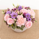 Arrangement floral Mélia rond avec roses roses, lisianthus violets, œillets et wax violet dans contenant blanc